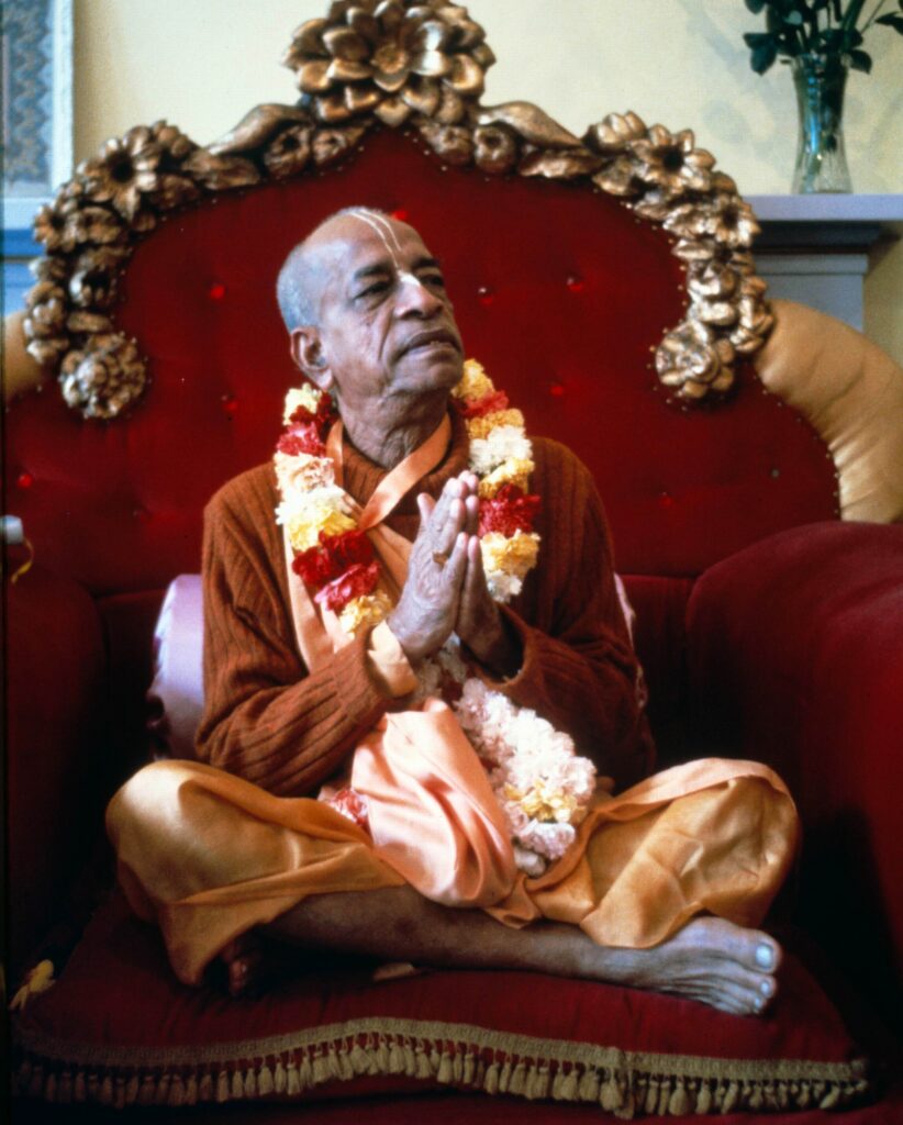 Srila Prabhupada sitting on a vyasasana (rasied seat), with folded hands. He is wearing a garland and looking up thoughtfully.
