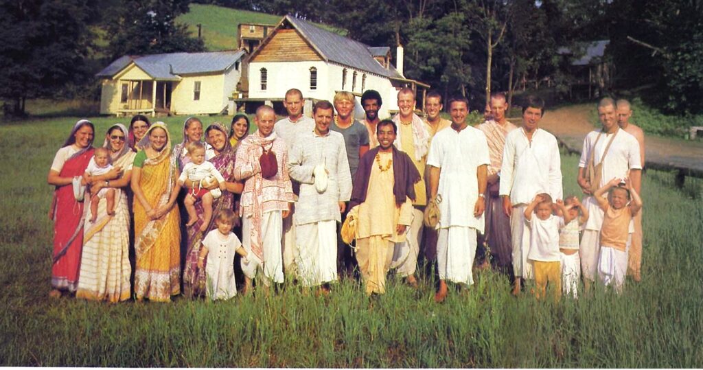 Vintage photo from the 1970s of about 20 devotees of various ages standing outside in the grass in front of farm buildings.