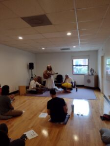 3 musicians performing kirtan while several participants sit on a wooden floor.