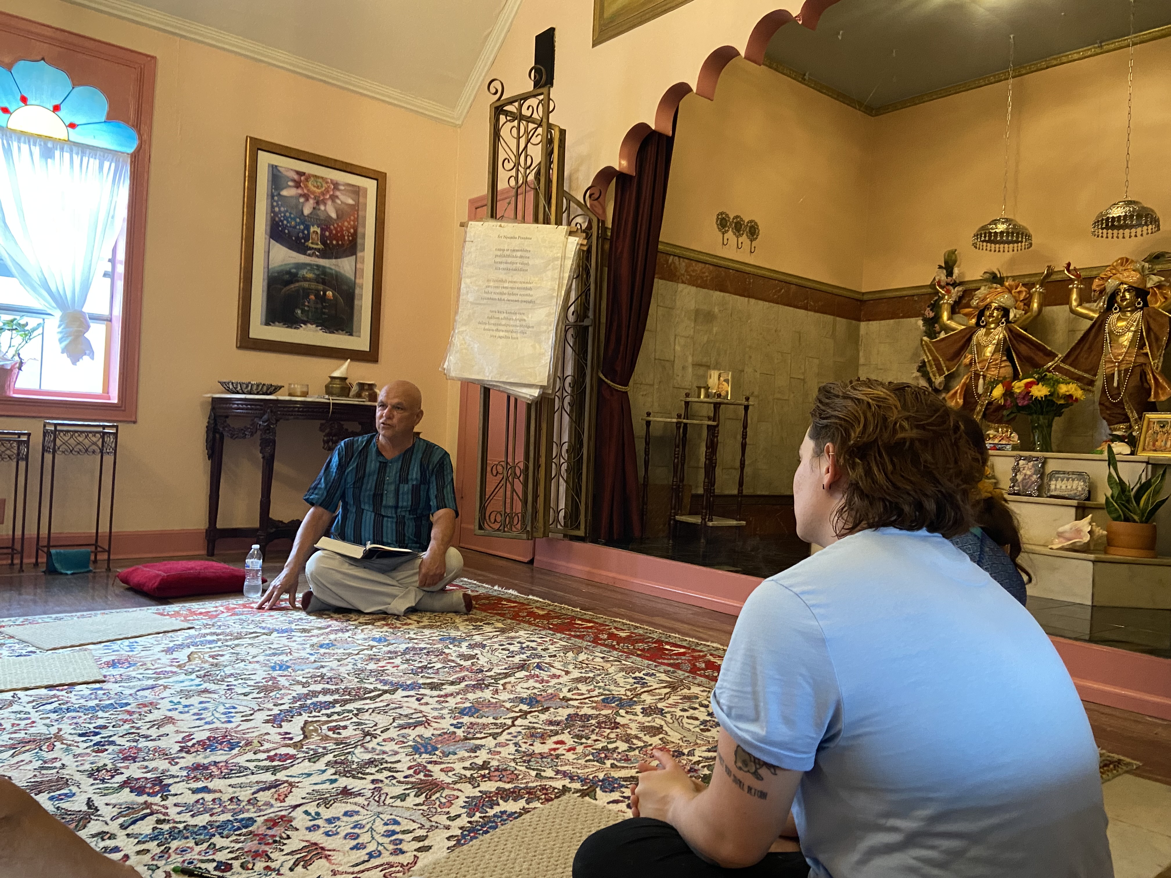 A devotee seated on the floor with a book open on his lap, while another devotee watches.