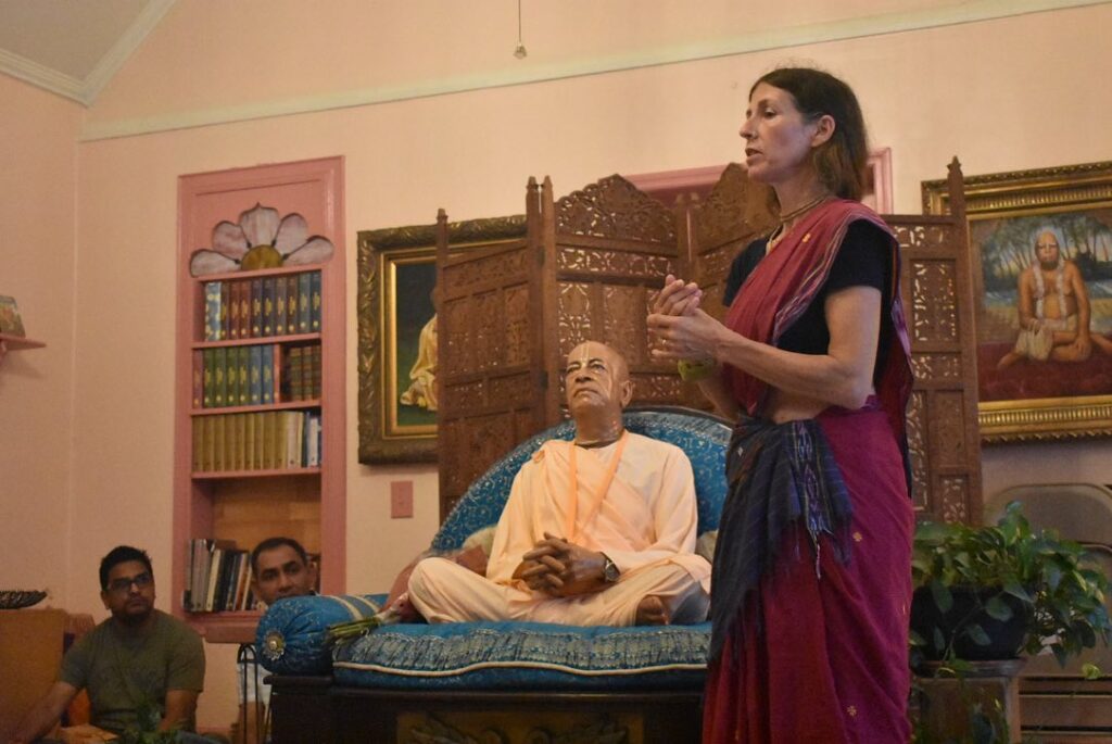 A woman standing in front of murti of Srila Prabhupada. A portrait of Bhaktivinoda Thakur hangs on the wall on one side.